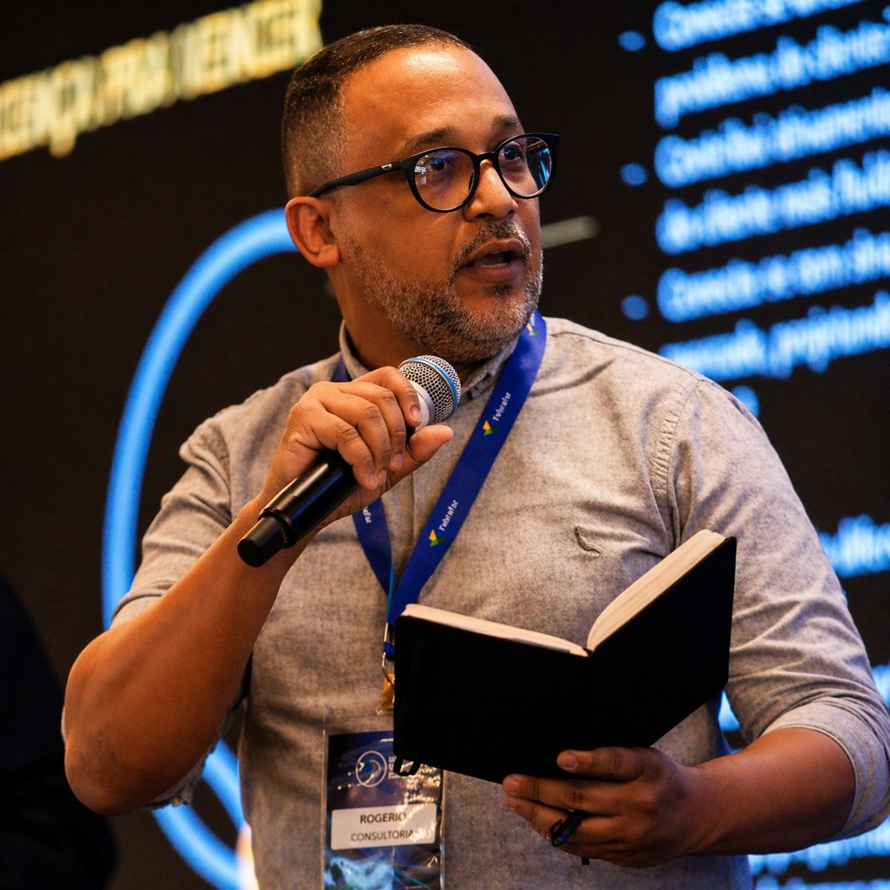 Rogerio Antunes fala ao microfone em um evento, com um caderno aberto na mão.